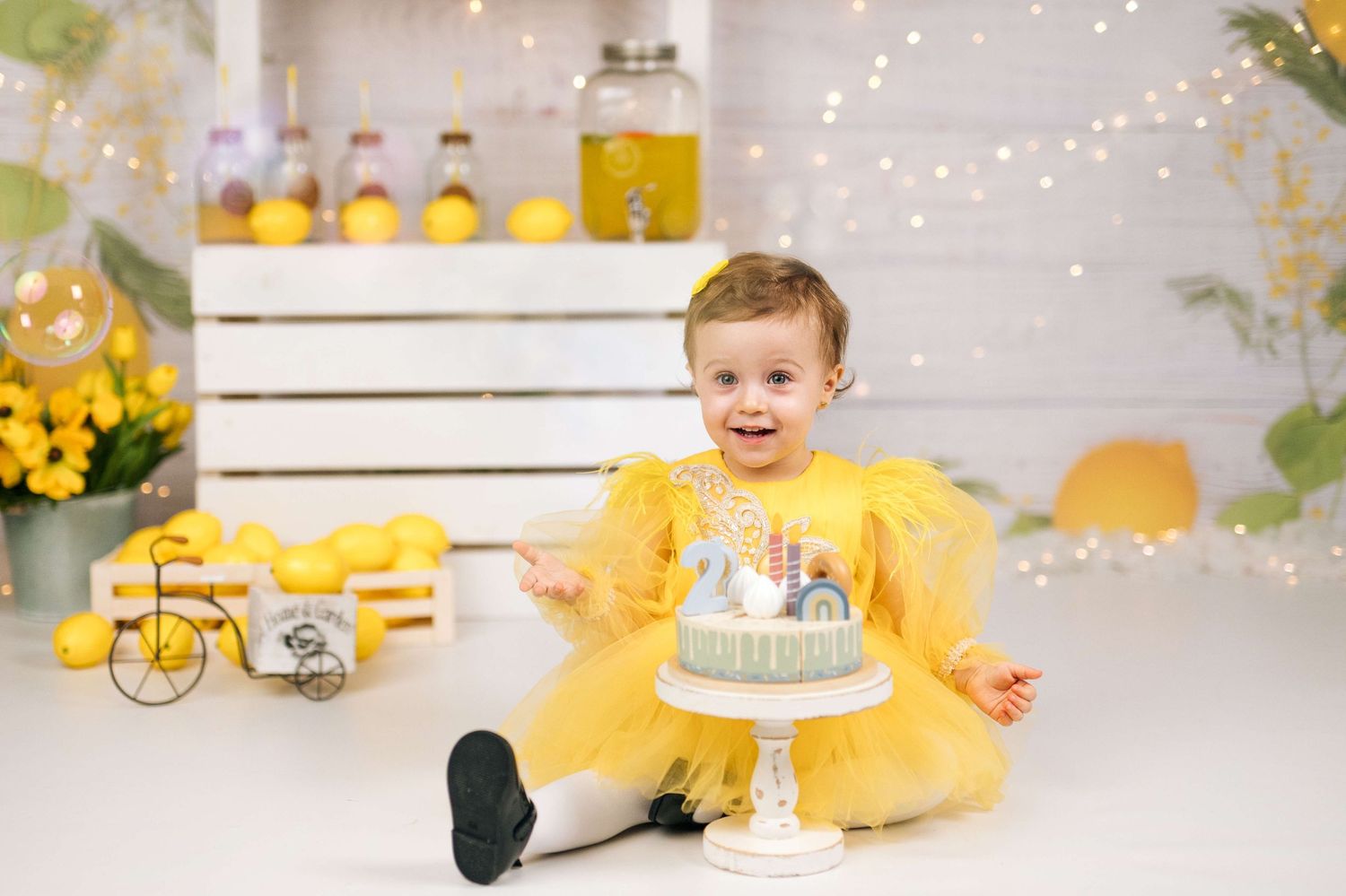A smiling baby girl in a yellow dress sits beside a birthday cake in a bright yellow-themed first birthday photo setup.