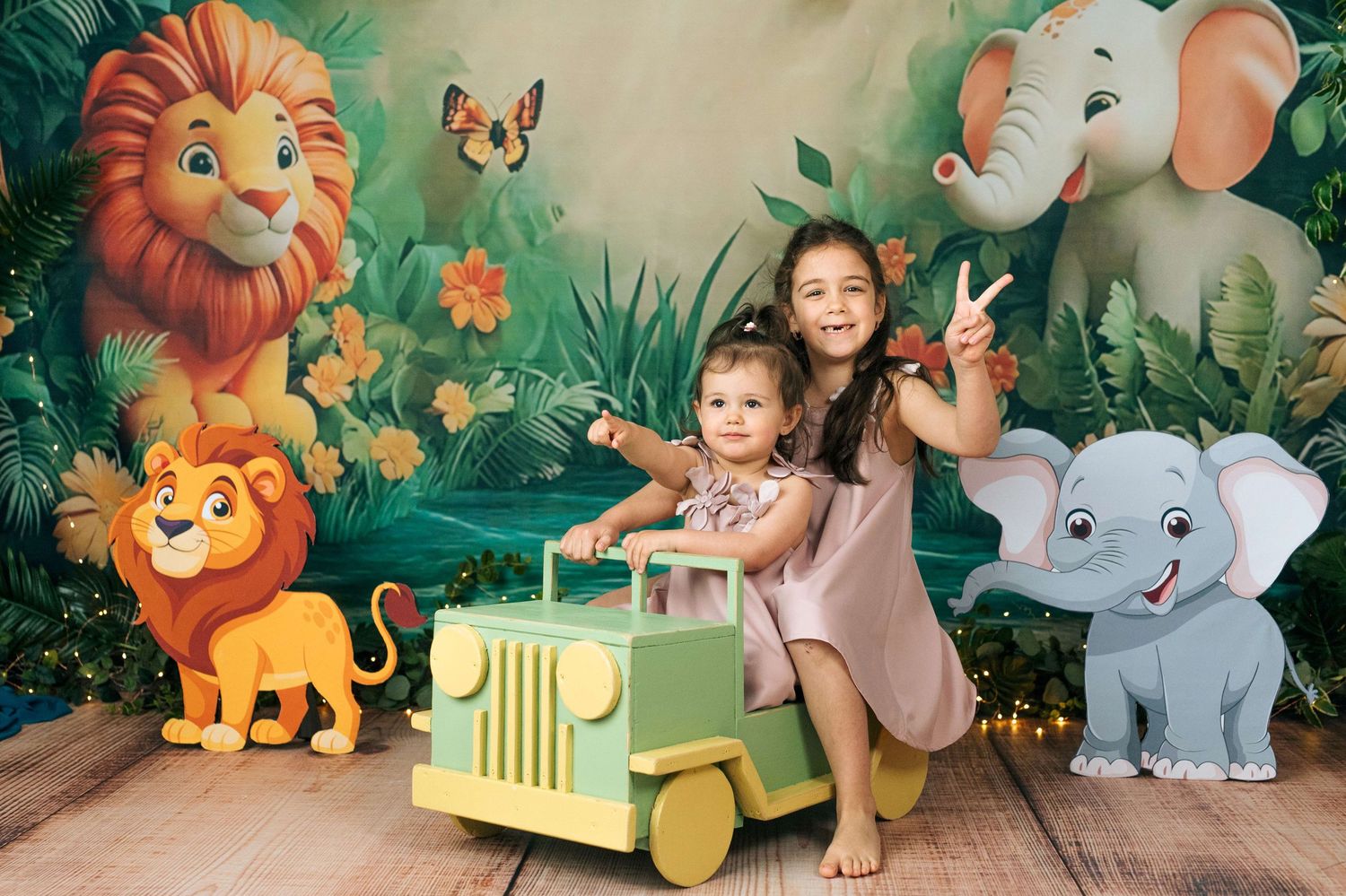 Two young girls pose on a toy jeep in front of a vibrant jungle backdrop featuring cartoon lions and elephants.