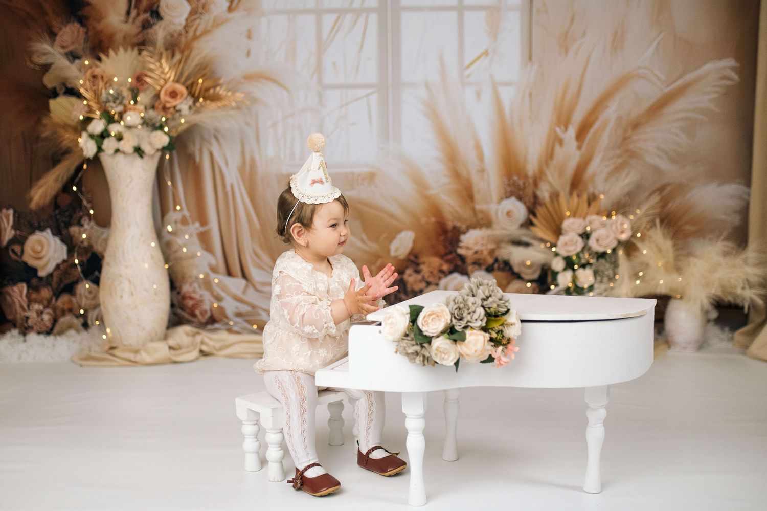 Toddler in white outfit and birthday hat clapping hands beside a white mini piano with floral boho decor.