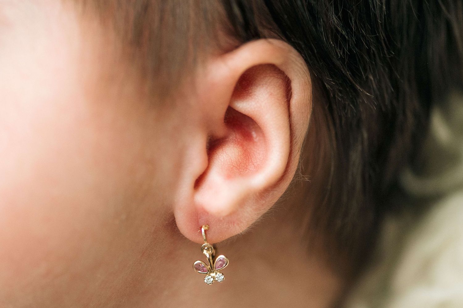 Close-up of a child's ear wearing a delicate gold fleur-de-lis drop earring with small gemstones.