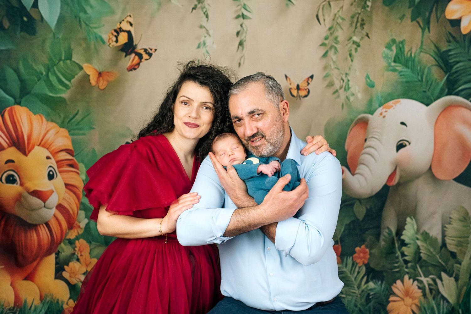 Family newborn photo shoot with jungle backdrop featuring parents in red and blue holding sleeping baby.