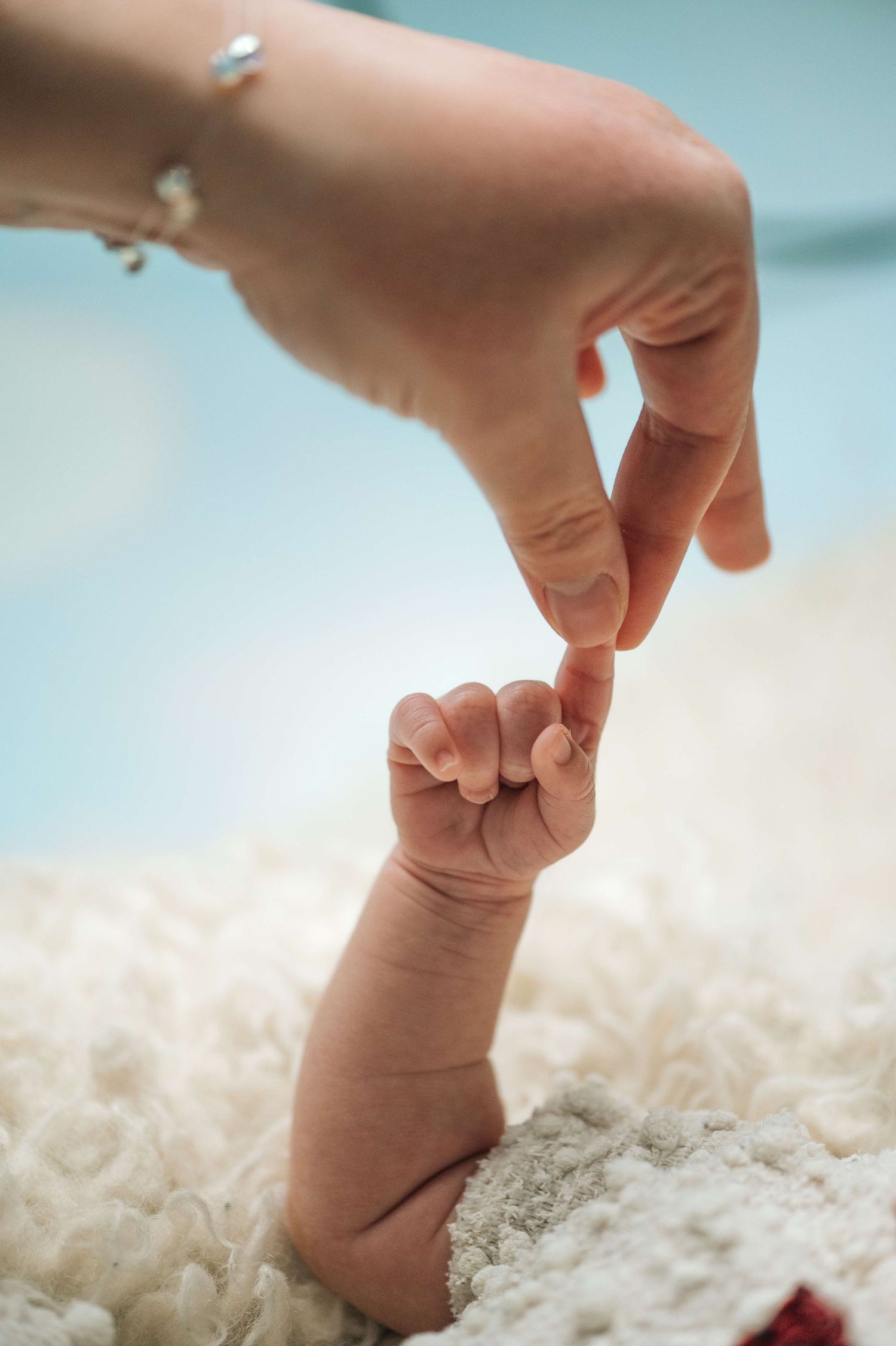 A newborn baby's tiny hand grasping an adult's finger on a soft white blanket.