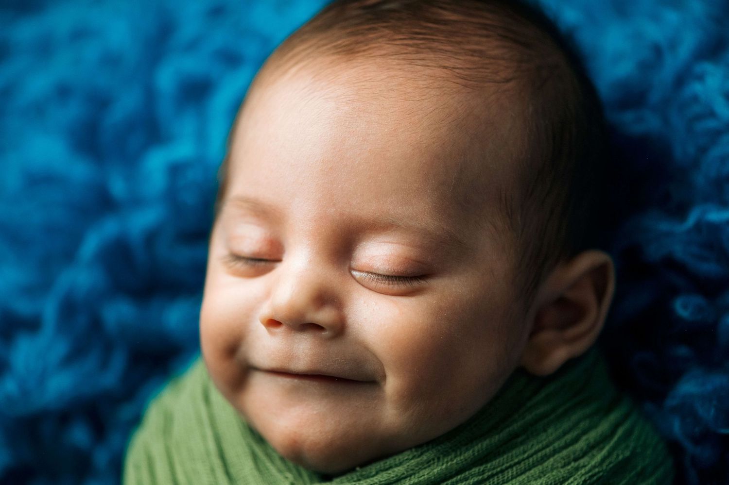Peaceful newborn baby smiling while sleeping, swaddled in green wrap on a soft blue textured blanket.