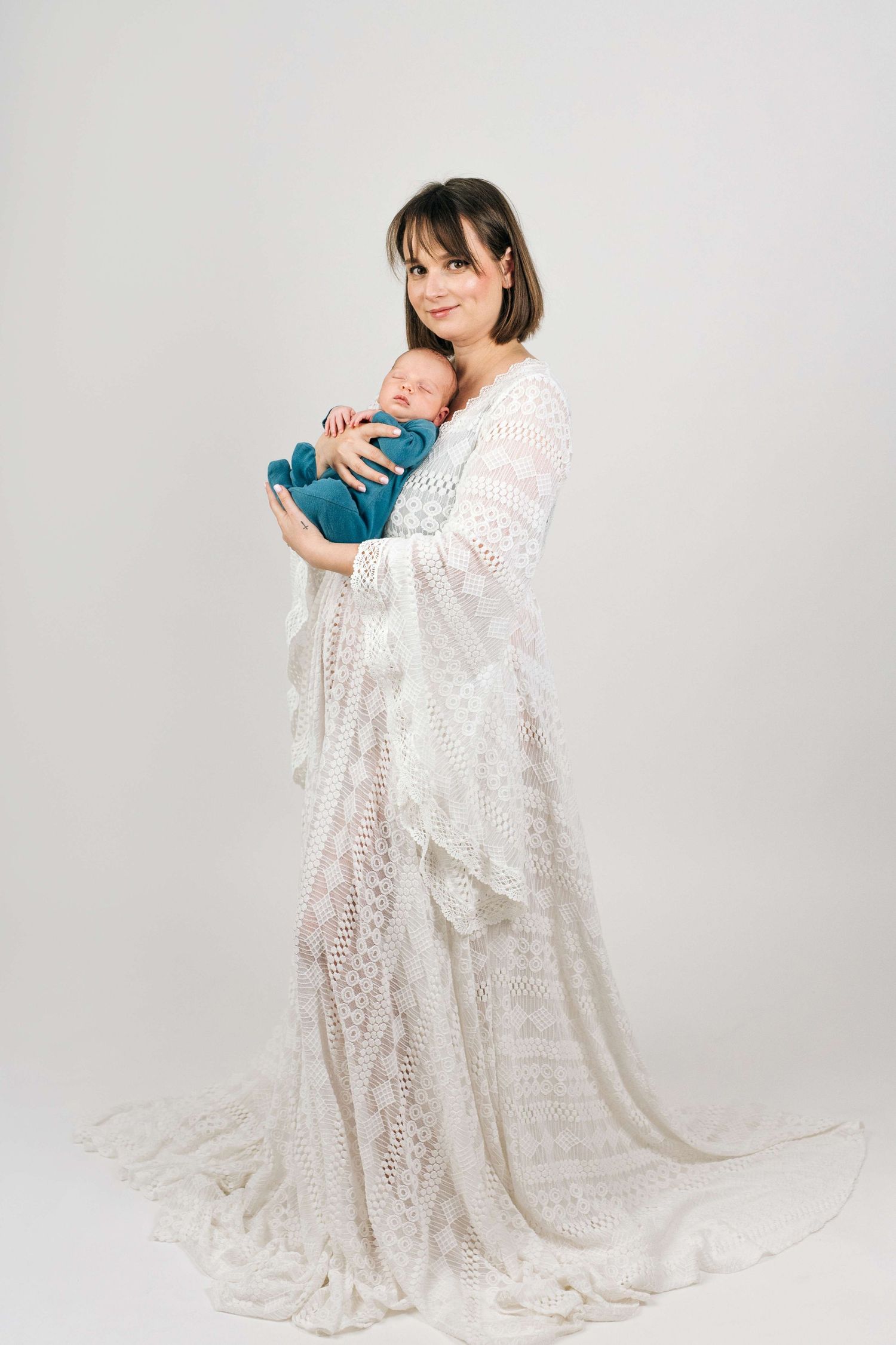 A mother in a white lace gown holds her sleeping newborn wrapped in teal fabric against a white background.