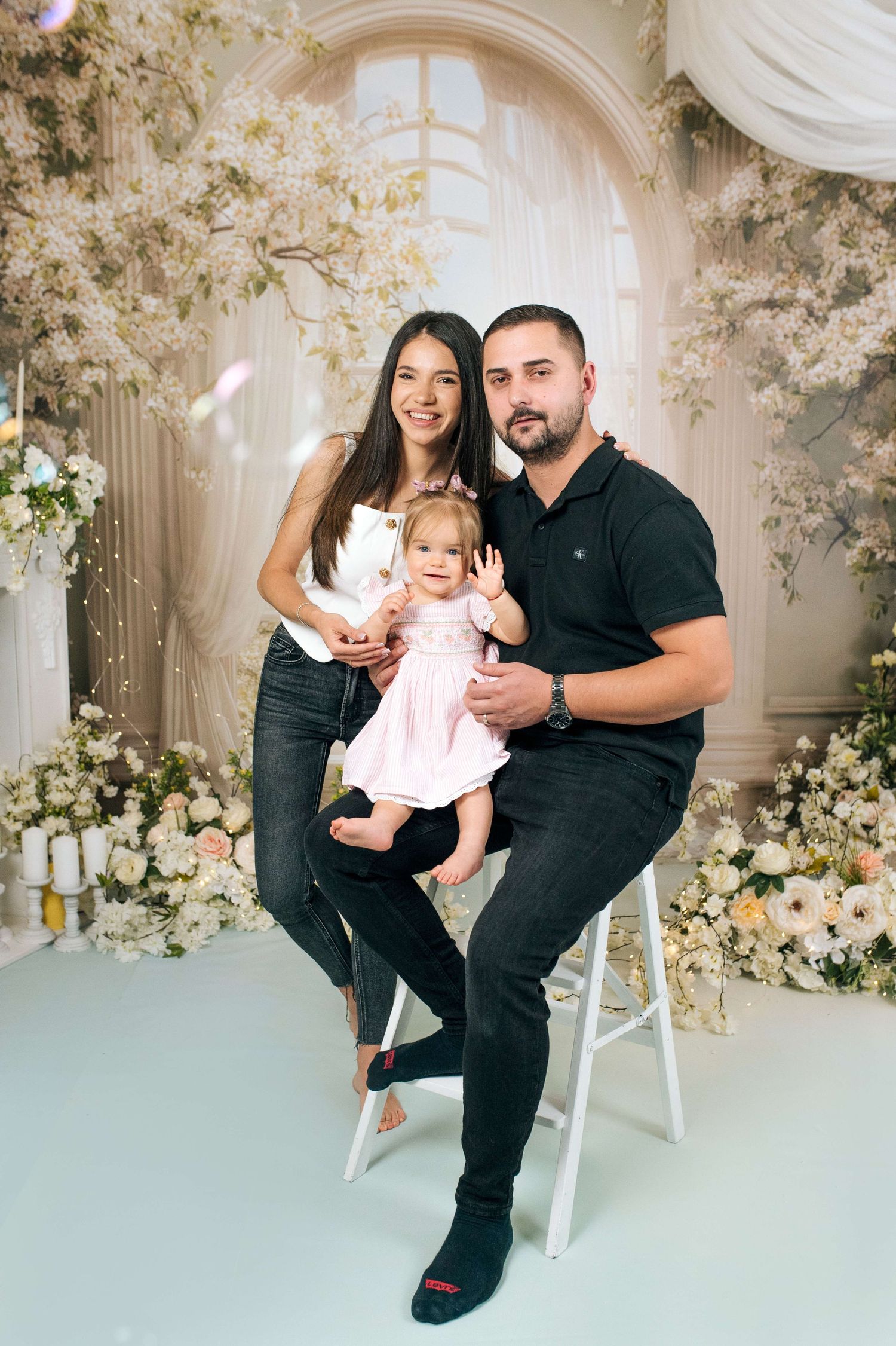 Young family with baby girl poses in floral studio setting, parents in black outfits with toddler in pink dress.
