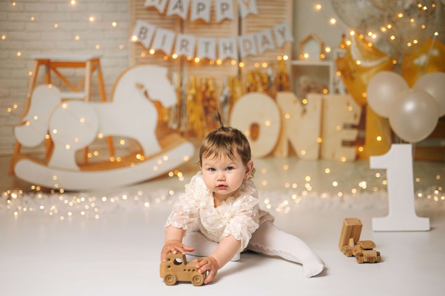 Baby girl in white dress plays with toy car at first birthday party with ONE sign and fairy lights.