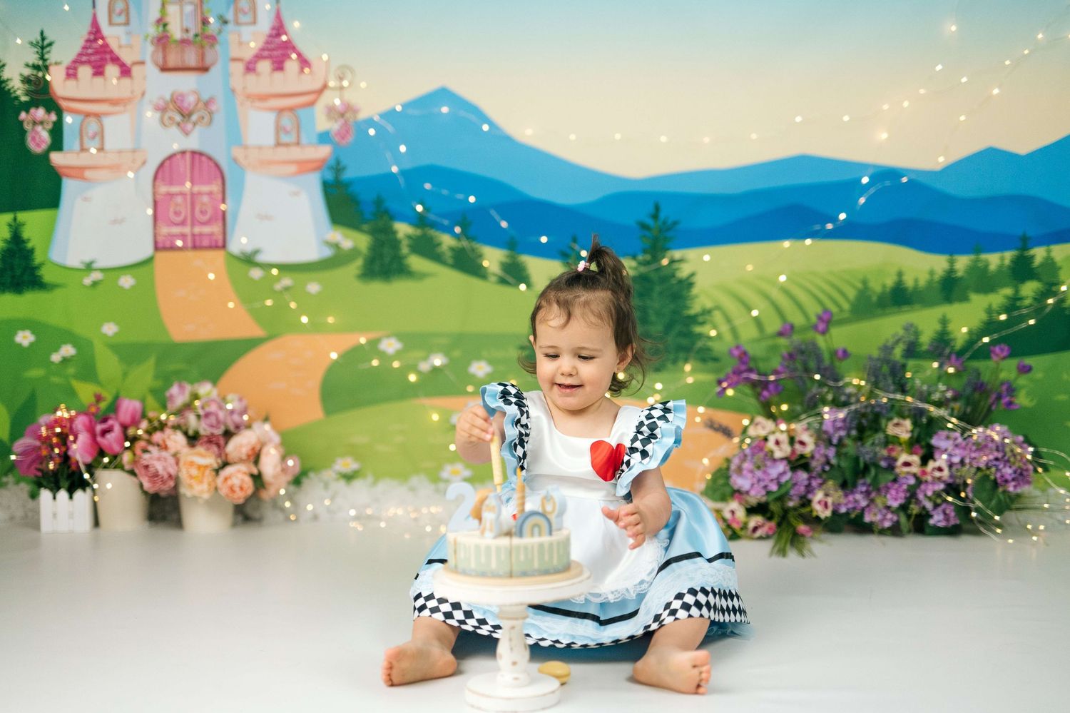 Toddler girl in blue dress celebrates 1st birthday with cake against a fairy tale castle backdrop with flowers.