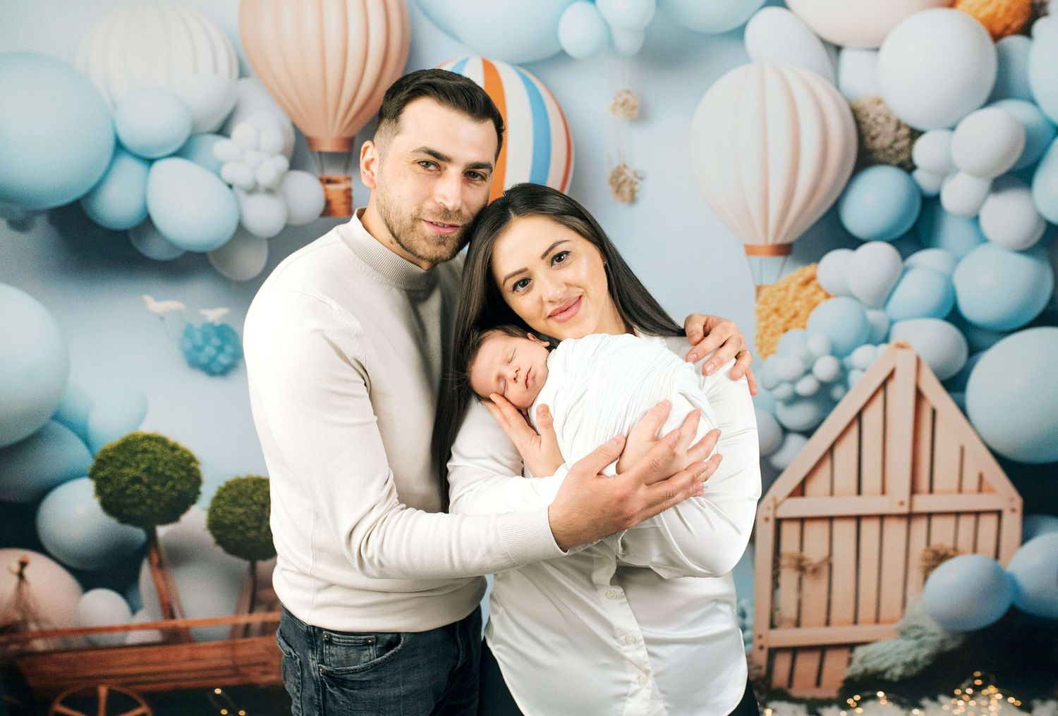 A couple holds their newborn baby in front of a blue balloon backdrop with hot air balloon decorations.