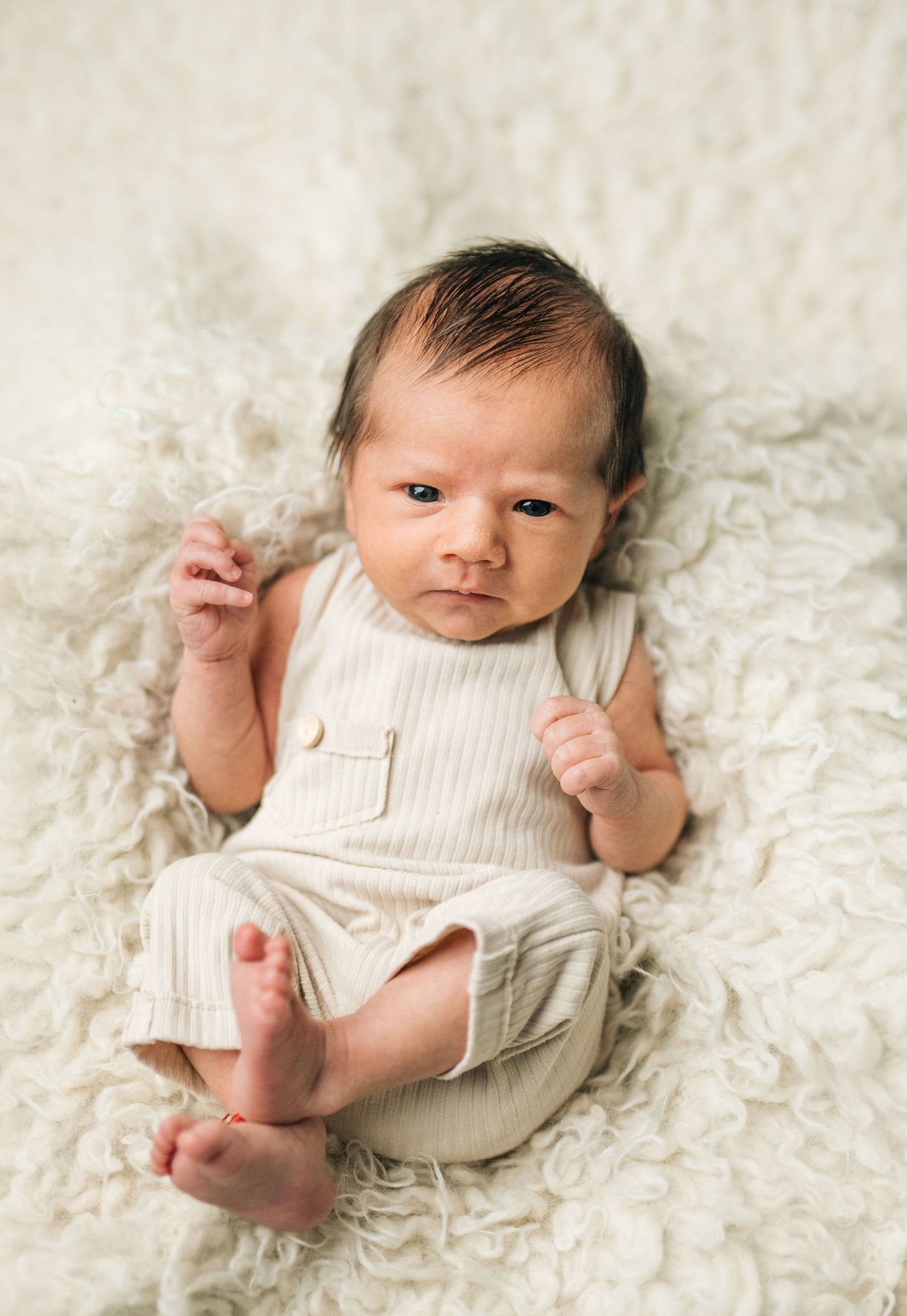 Newborn baby in a cream ribbed romper lying on a fluffy white blanket with fists raised, looking alert.