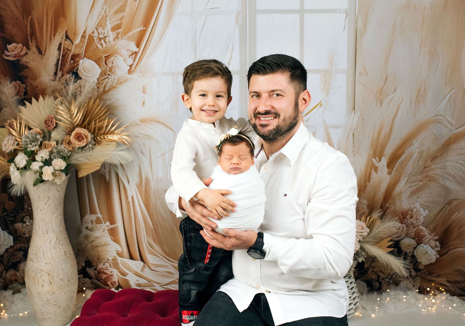 Father smiles holding newborn baby while young son poses beside him in a warm, floral studio portrait.