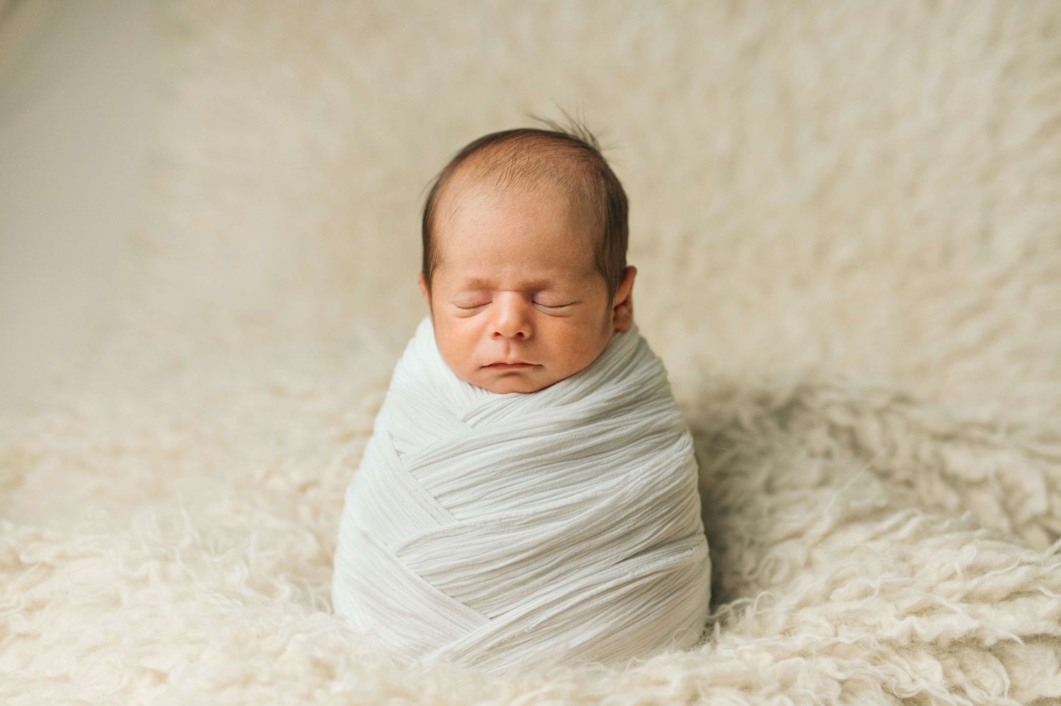 Newborn baby swaddled in white wrap sleeping peacefully on fluffy cream blanket in professional studio photo.