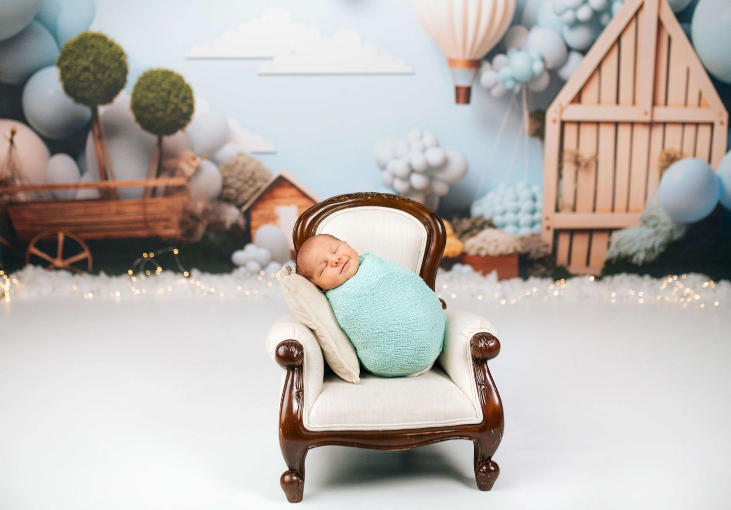 Newborn baby wrapped in teal swaddle sleeping on miniature armchair with hot air balloon backdrop.