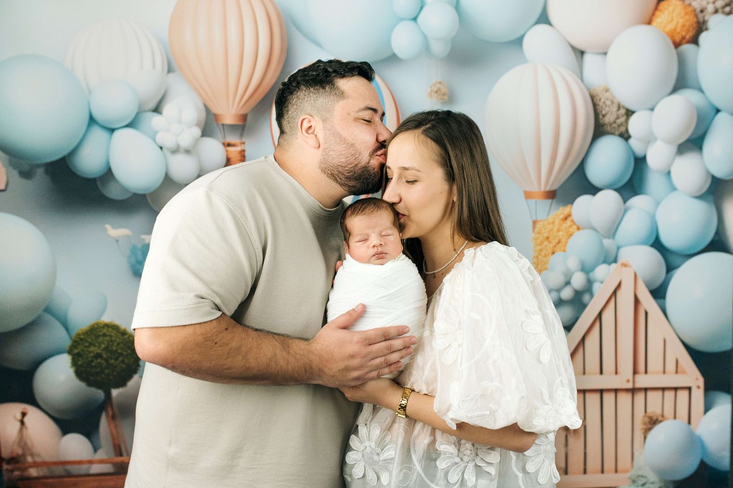 Parents kissing their newborn baby during a professional photoshoot with blue balloon and hot air balloon backdrop.