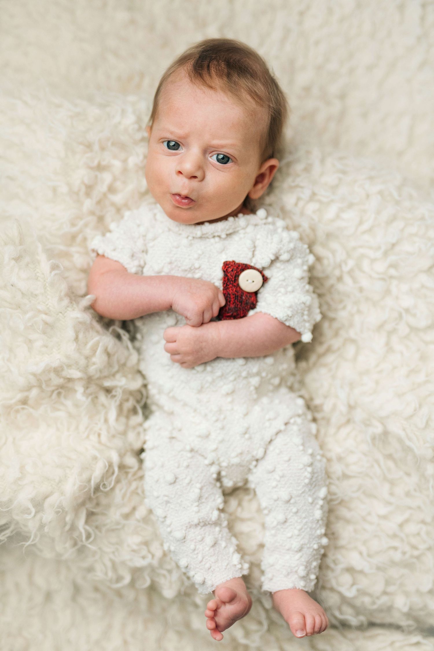 Newborn baby in white fluffy outfit lying on cream fur blanket, holding a small red knitted toy.