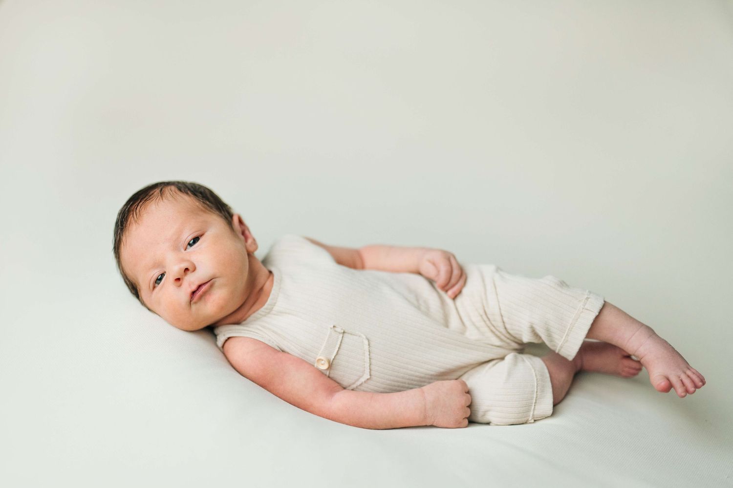 Newborn baby in cream onesie lying on white backdrop in professional studio portrait photography.