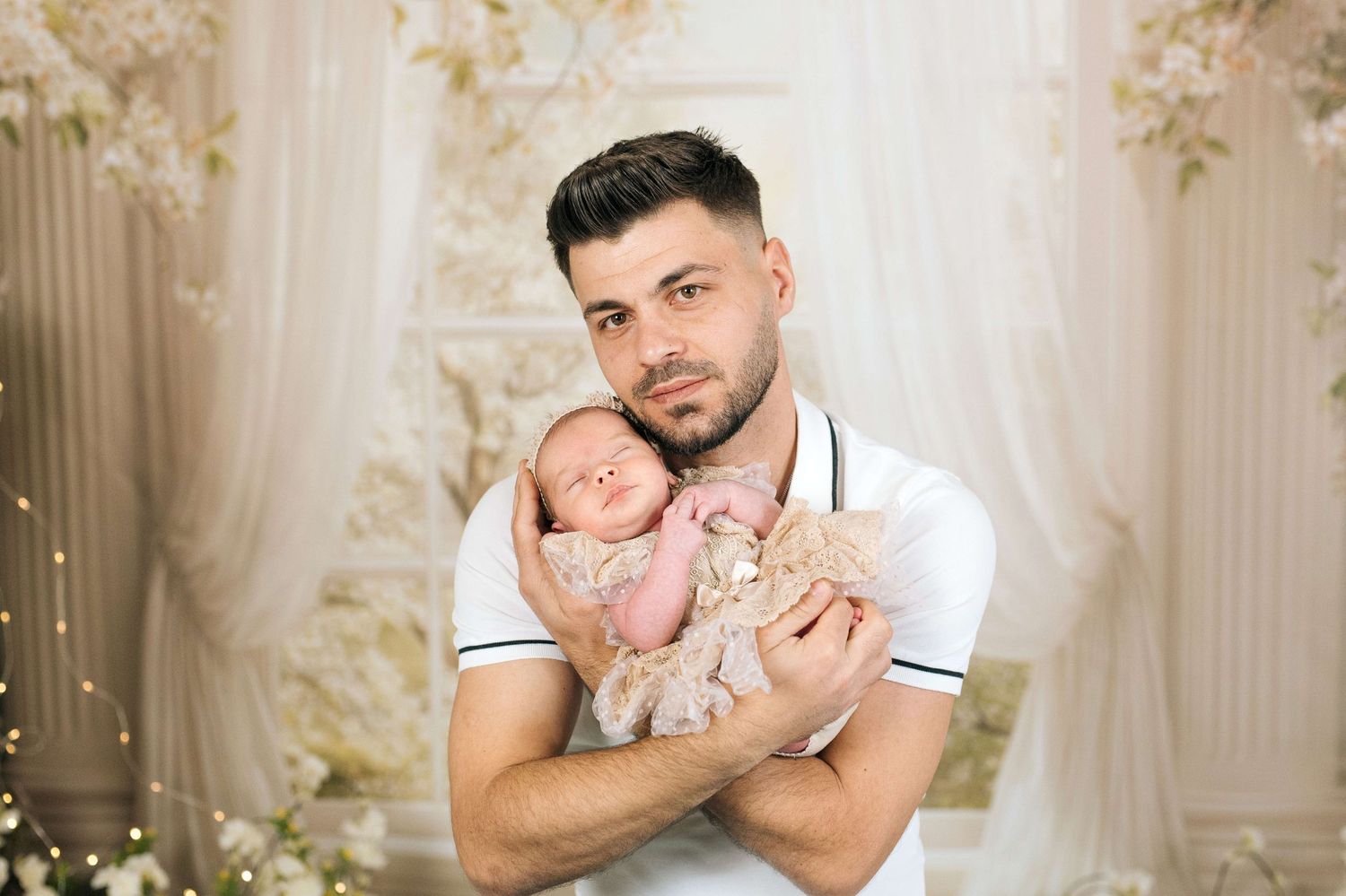 A young father with dark hair and beard tenderly holds his sleeping newborn baby in a floral studio setting.