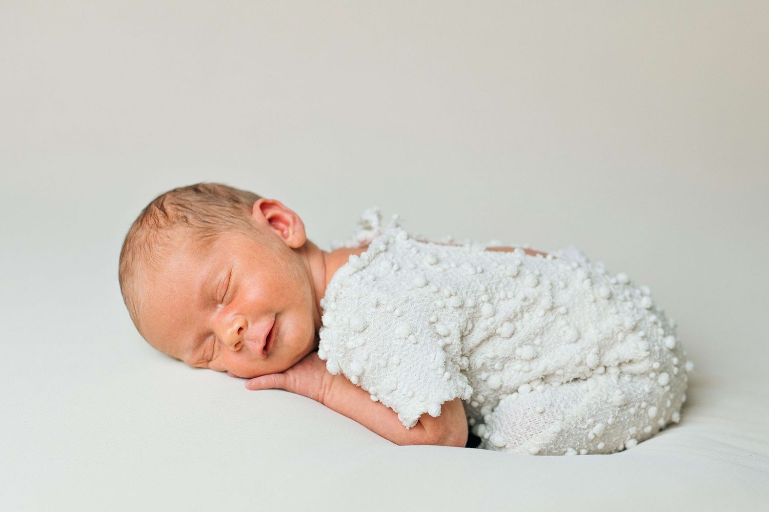 Peaceful newborn baby sleeping wrapped in white textured blanket on soft white background.