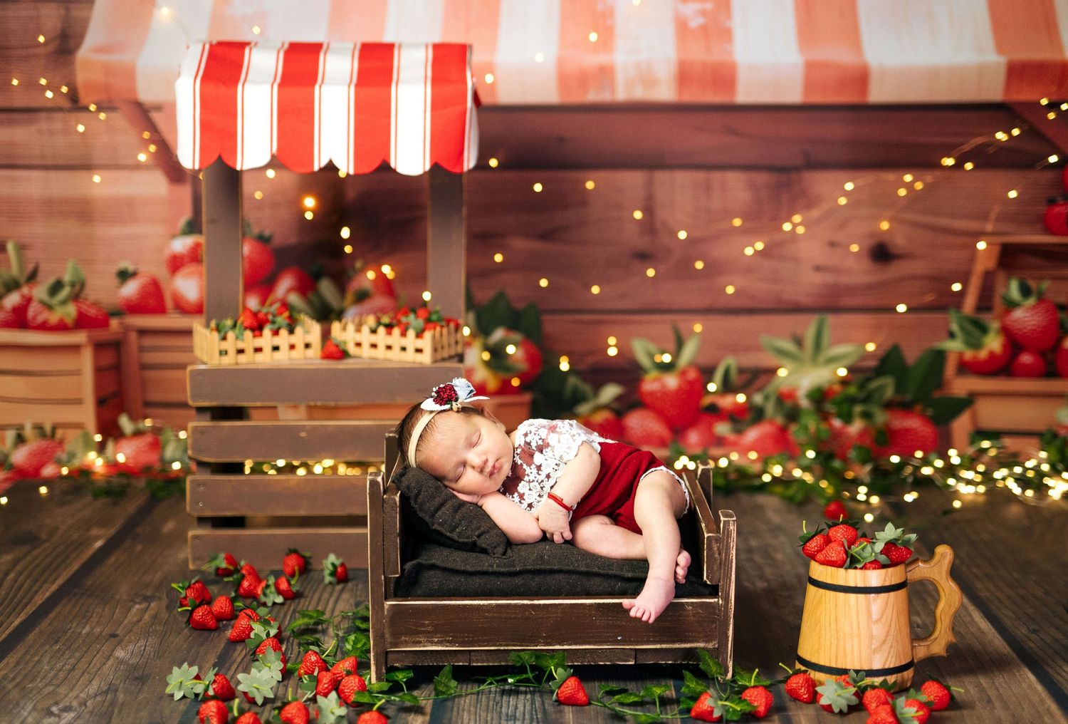 Newborn baby sleeping in a wooden crate surrounded by strawberries and fairy lights in a charming fruit stand setting.