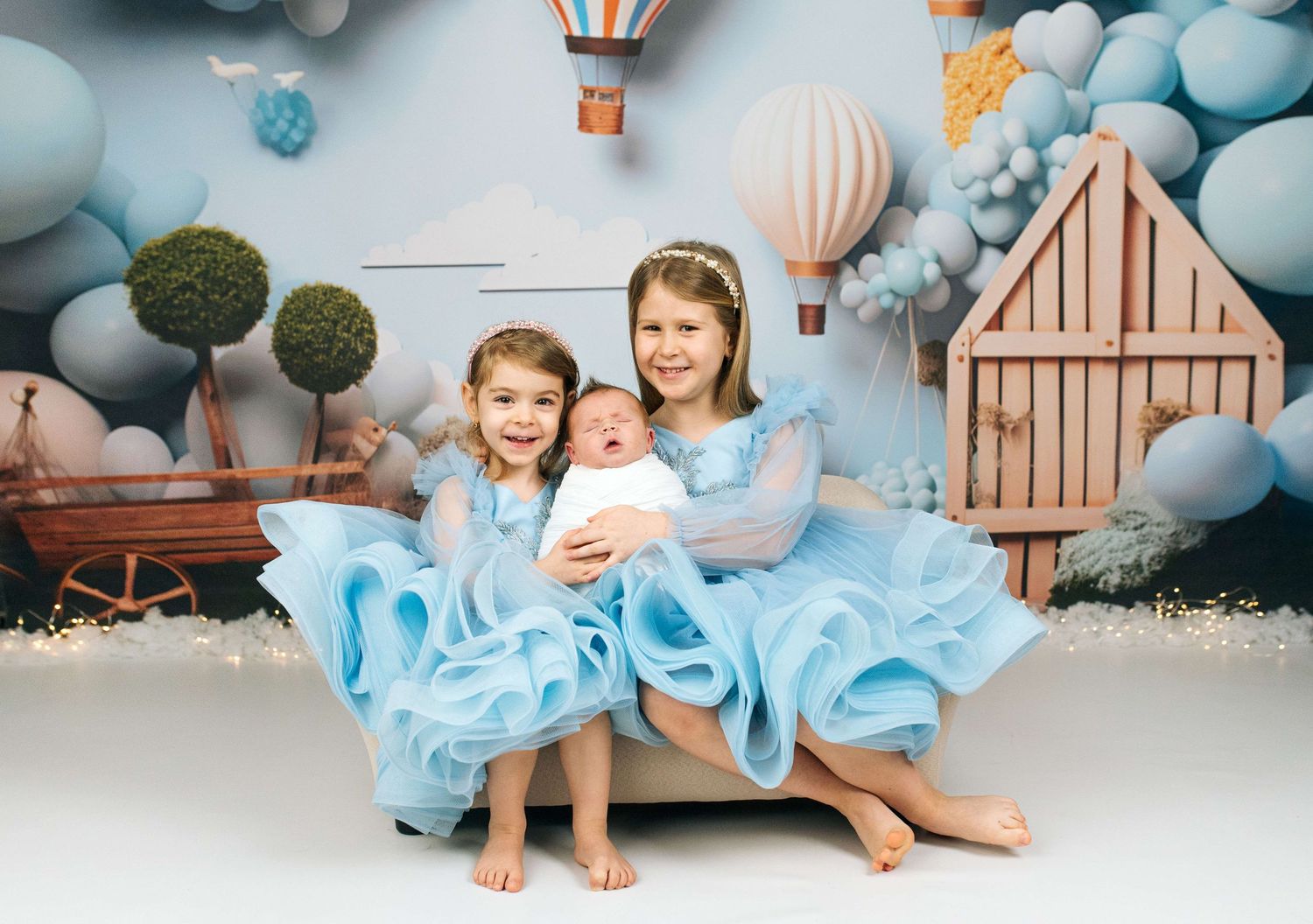 Two girls in blue tulle dresses pose with a newborn baby in a whimsical hot air balloon themed photo studio.