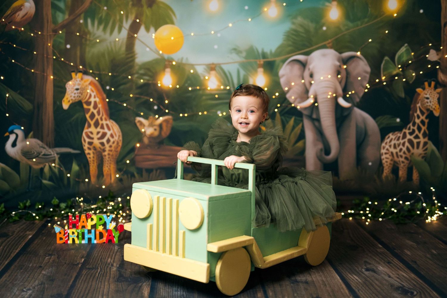 Toddler in green dress sitting in toy jeep for birthday photos with jungle safari backdrop featuring elephants and giraffes.