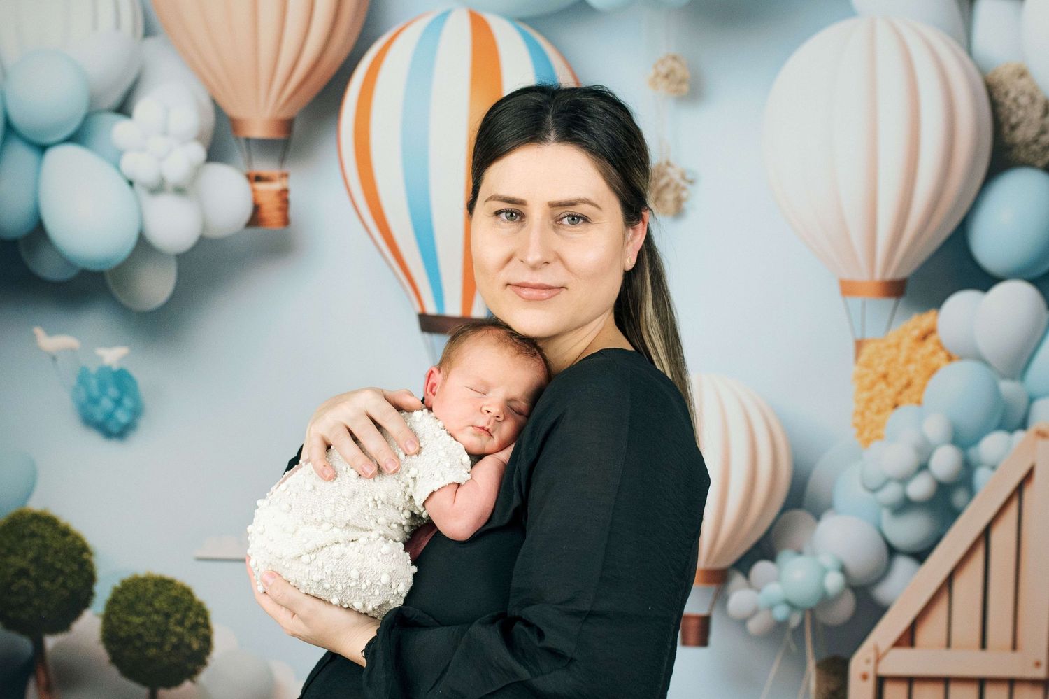 A mother in black dress holds sleeping newborn baby against colorful hot air balloon nursery backdrop.