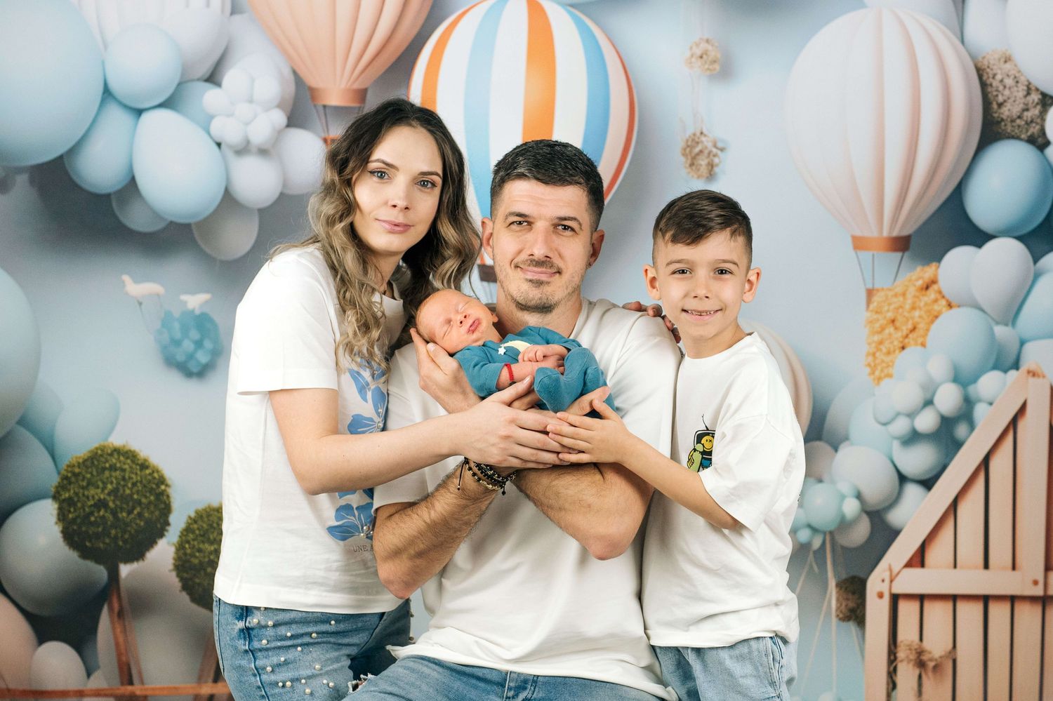 Family of four posing with newborn baby in front of hot air balloon backdrop in matching white outfits.