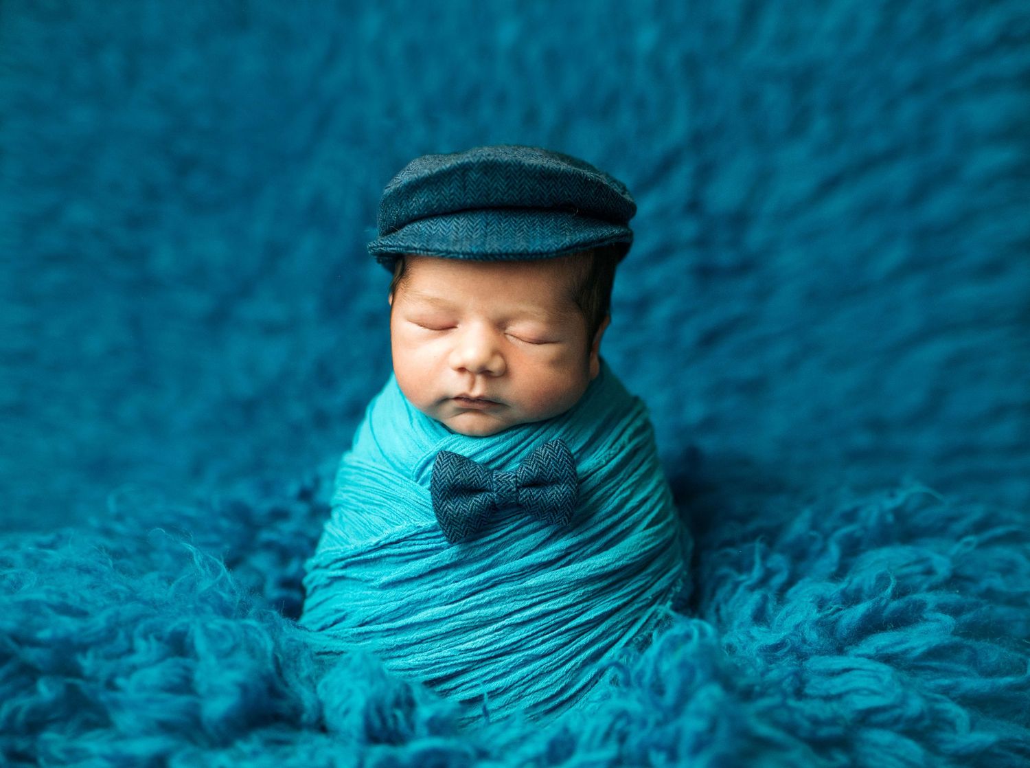 Adorable newborn baby swaddled in teal wrap, wearing a tiny newsboy cap and bow tie, sleeping on a fluffy blue rug.