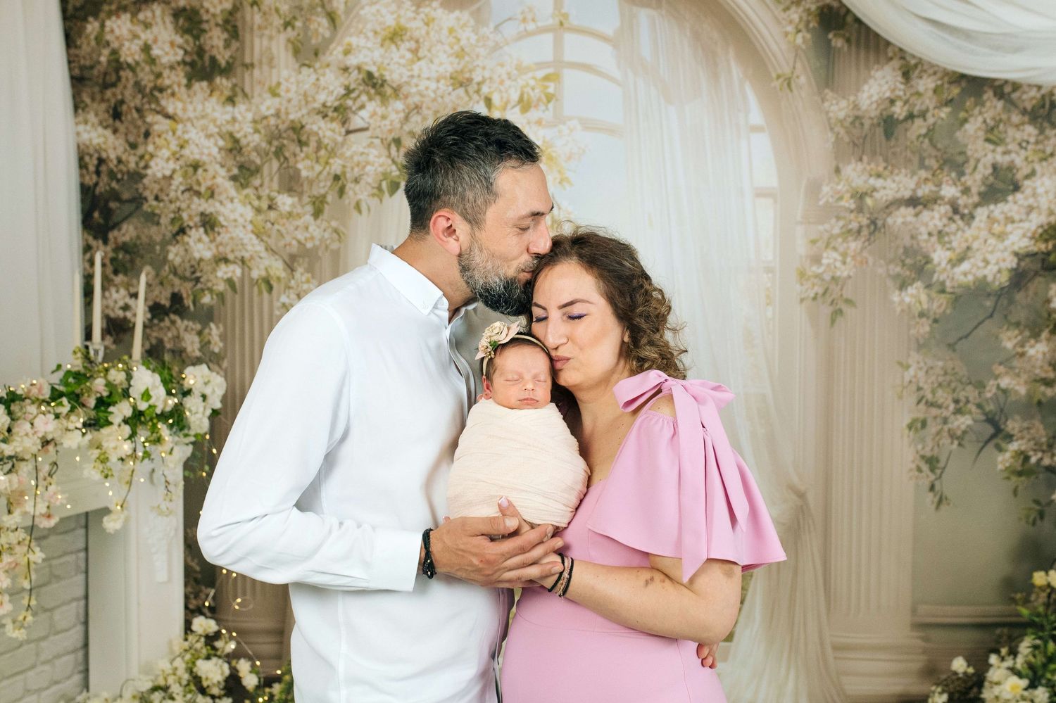 Parents tenderly kissing their swaddled newborn baby in elegant white floral studio photography setting.