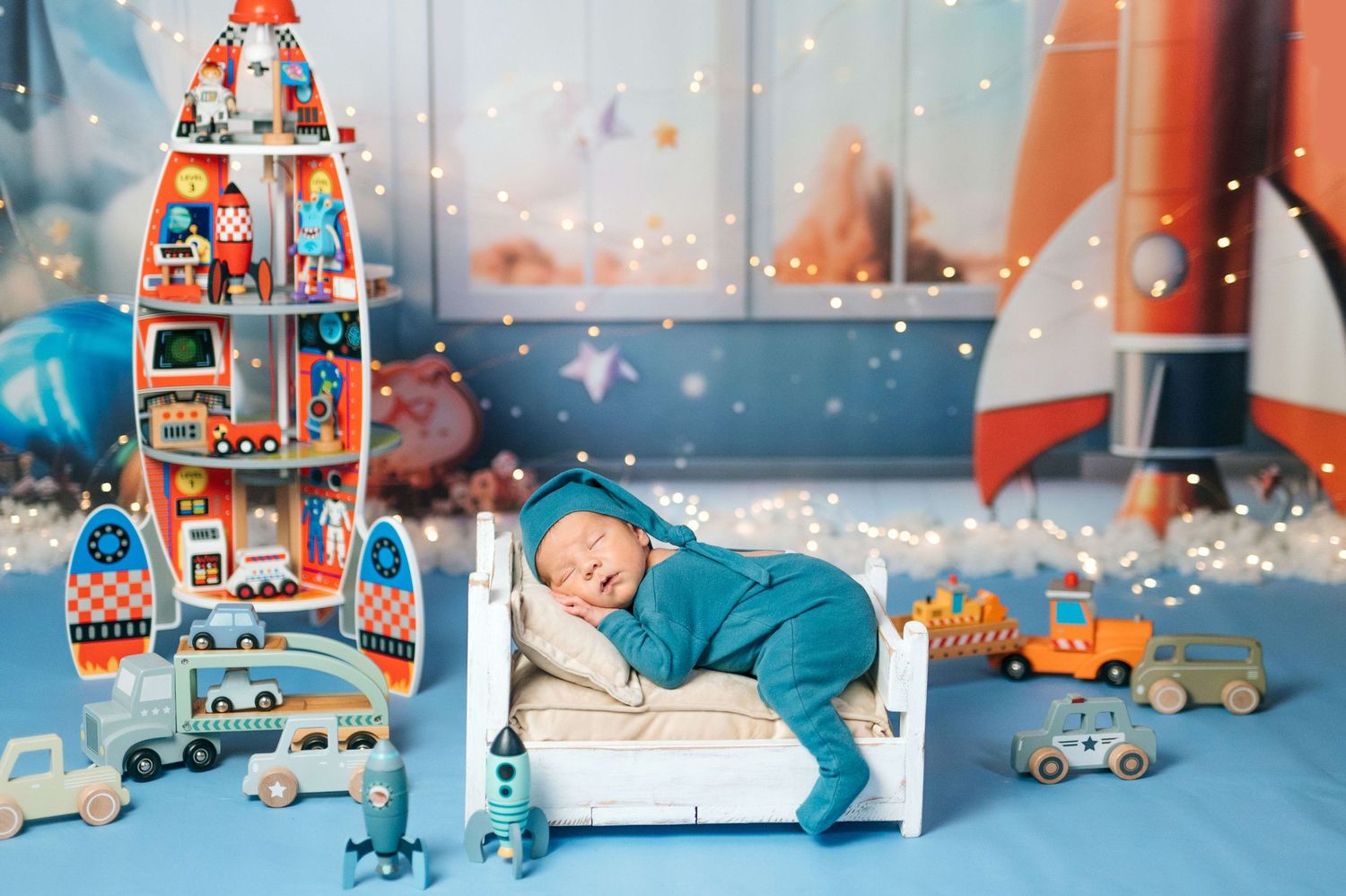 Newborn baby sleeping in tiny white bed surrounded by colorful toy rockets and cars in a space-themed nursery setting.