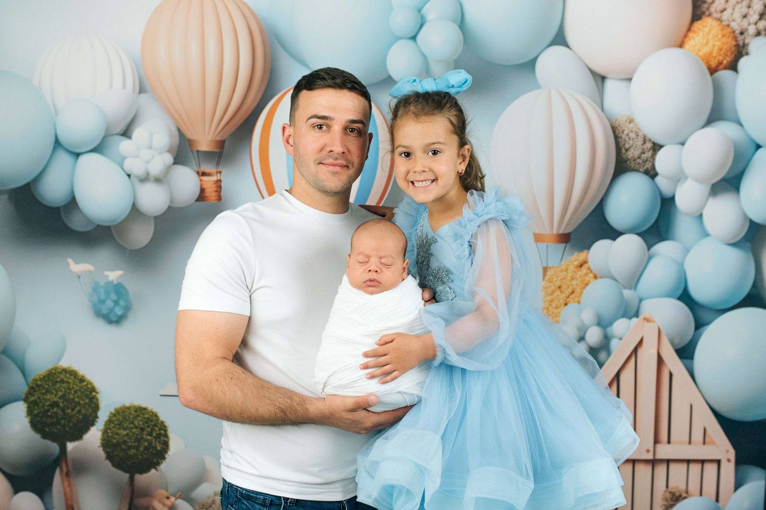 Father holds newborn baby while young girl in blue dress poses in front of balloon-filled backdrop.