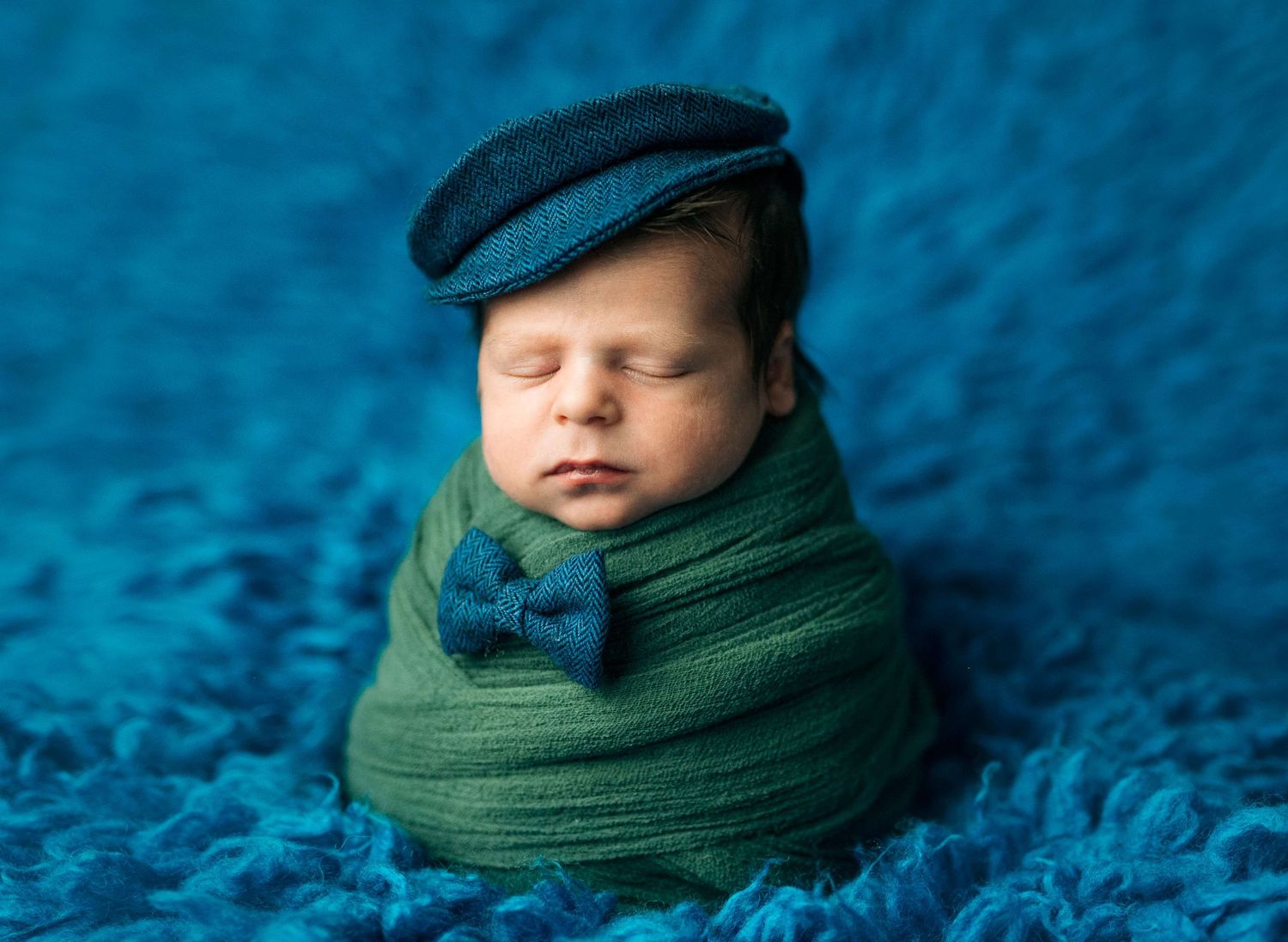 Newborn baby swaddled in green wrap wearing a blue flat cap and bow tie on a teal fluffy blanket.