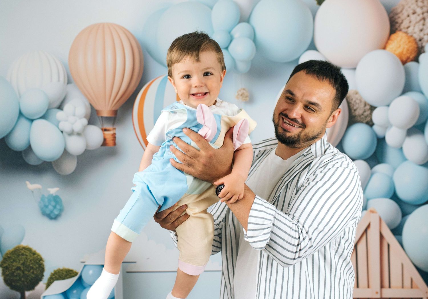 A smiling father holds his happy toddler son in a blue balloon-decorated studio photo shoot backdrop.