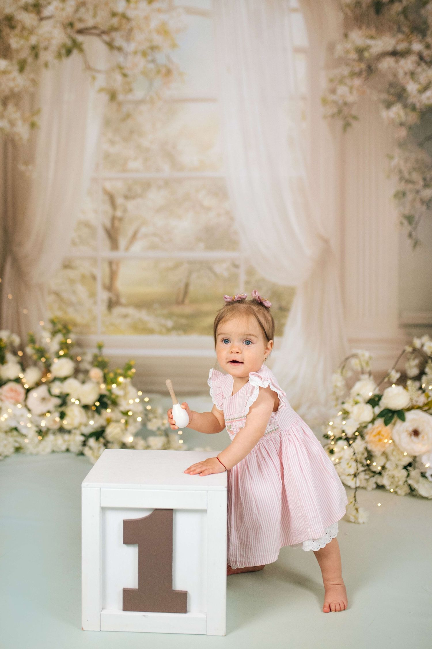 Baby girl in pink dress stands by a number 1 block, celebrating her first birthday with floral backdrop.