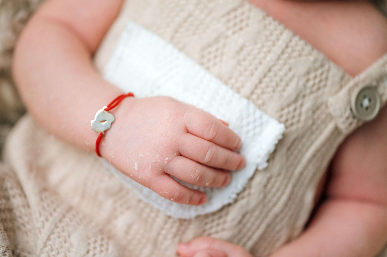 Close-up of a newborn baby's tiny hand wearing a red bracelet, dressed in a cozy beige knit outfit.