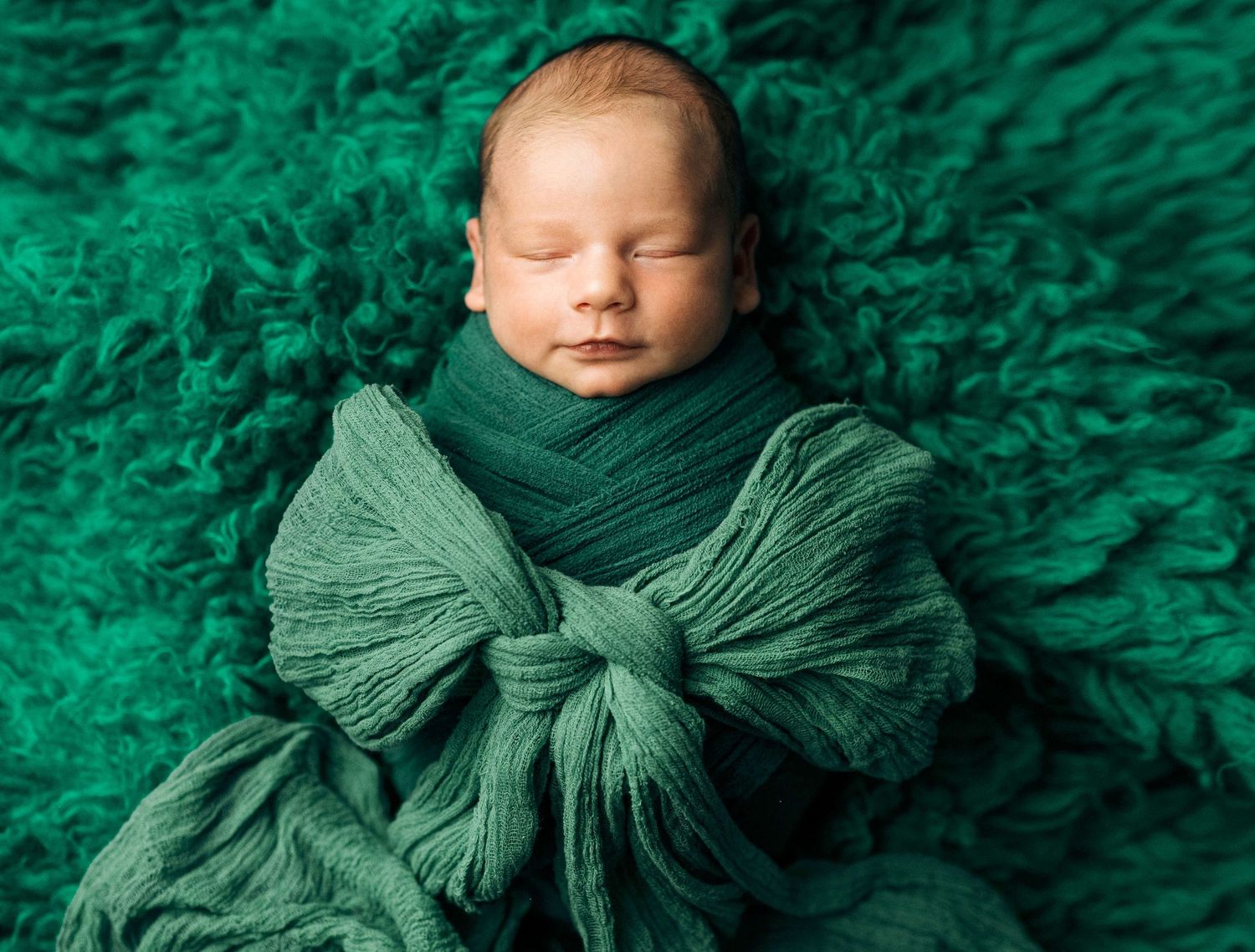Newborn baby sleeping peacefully wrapped in green fabric on a fluffy emerald green backdrop.