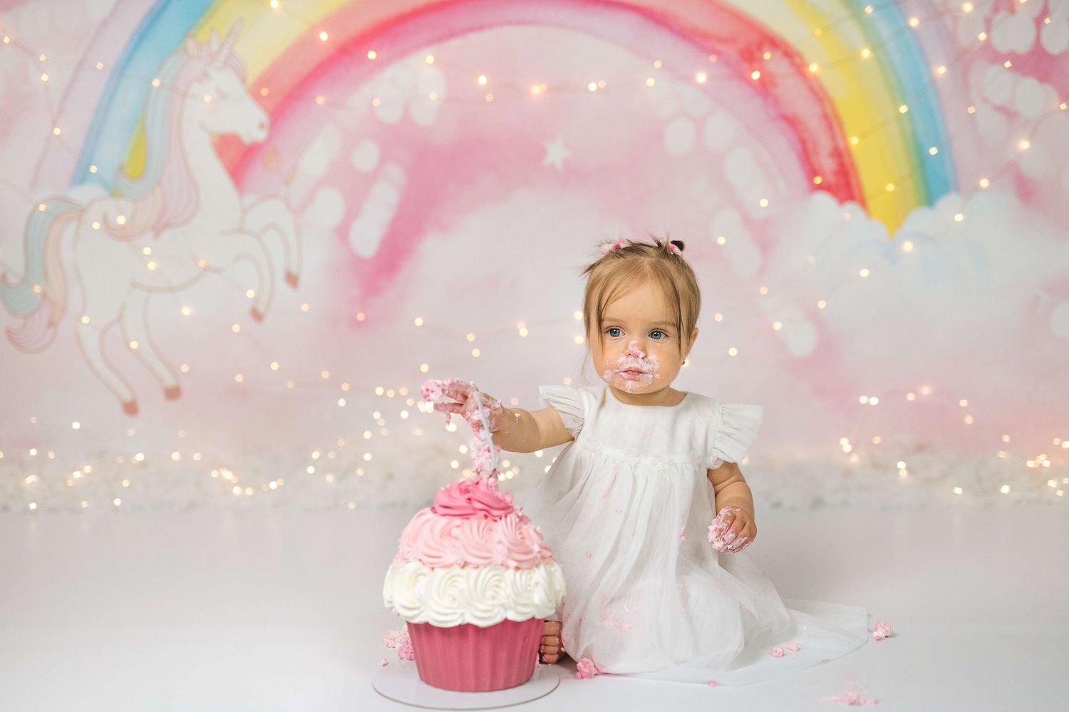 A baby girl in a white dress smashes a pink cupcake cake in front of a rainbow unicorn backdrop.