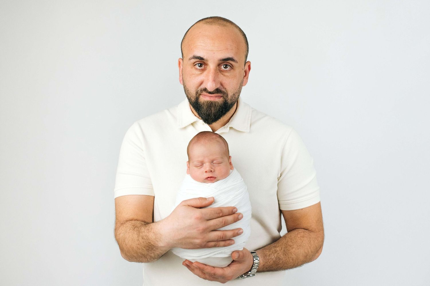 Proud bald bearded father holding sleeping newborn baby wrapped in white swaddle against light background.