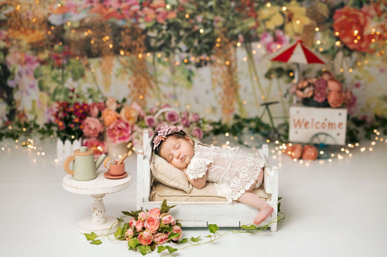Sleeping newborn baby girl in lace outfit on white miniature bed with floral backdrop and fairy lights.