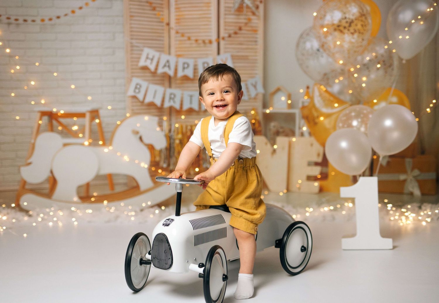 Baby boy smiles on toy car at first birthday photoshoot with balloons and Happy Birthday banner.