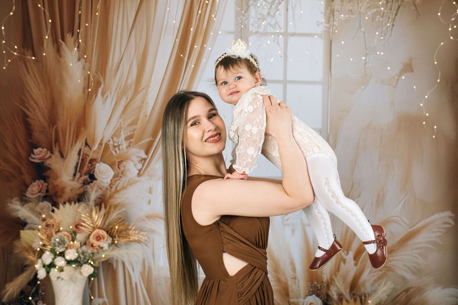 A smiling woman in a brown dress holds a baby girl in a white lace outfit in a boho-styled photo studio.