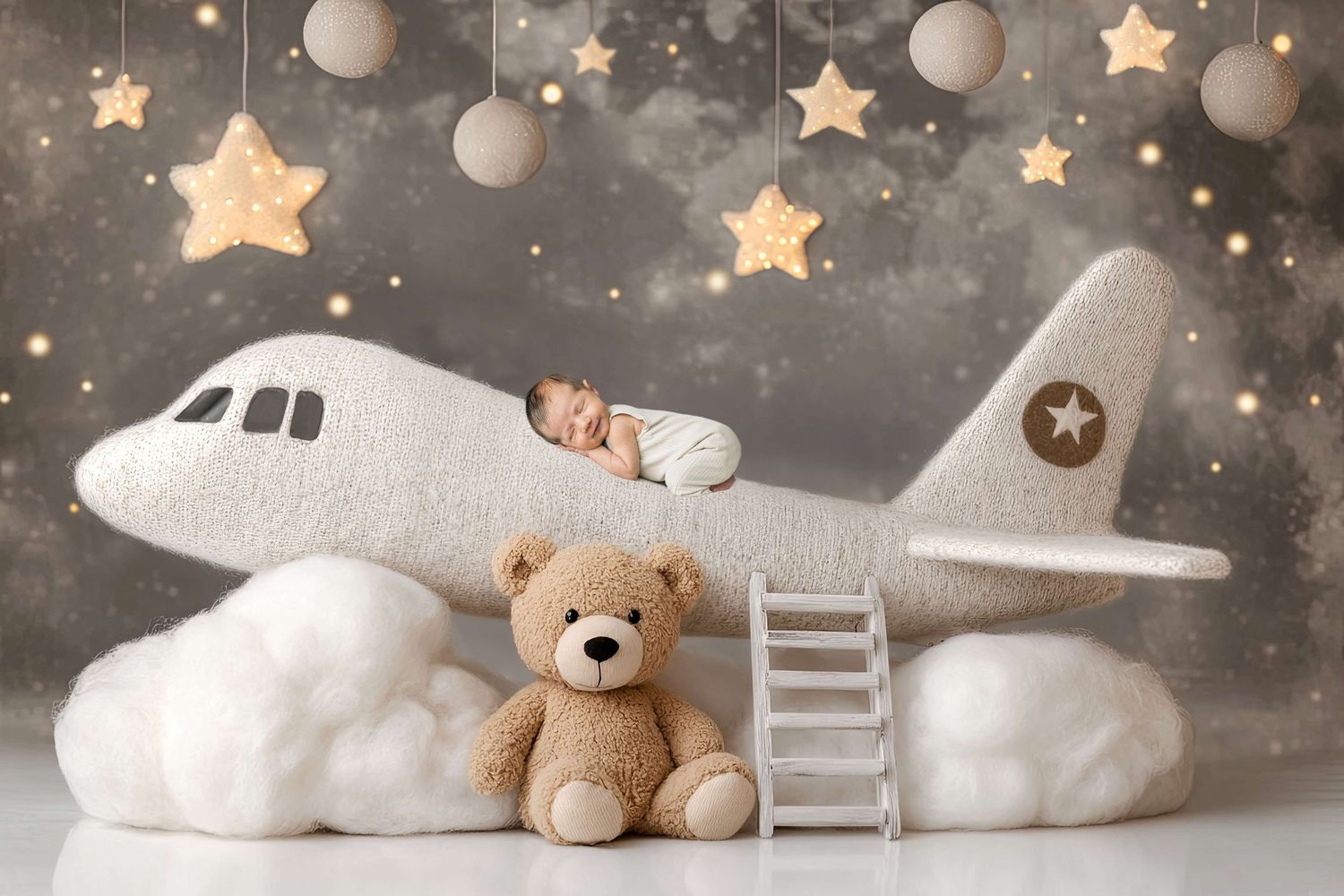 Newborn baby sleeping on a plush toy airplane with a teddy bear, stars, and clouds in a dreamy nursery photo.