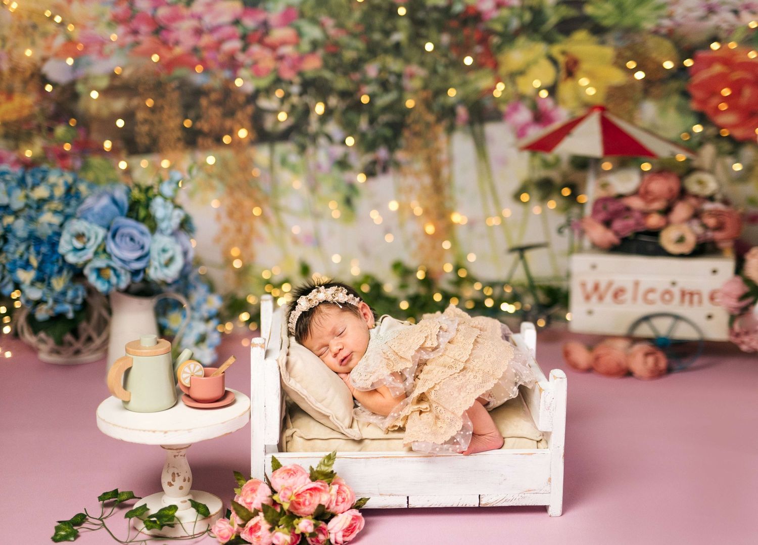 Newborn baby sleeping on a white miniature bed with floral wreath, surrounded by flowers and fairy lights in a dreamy studio setup.