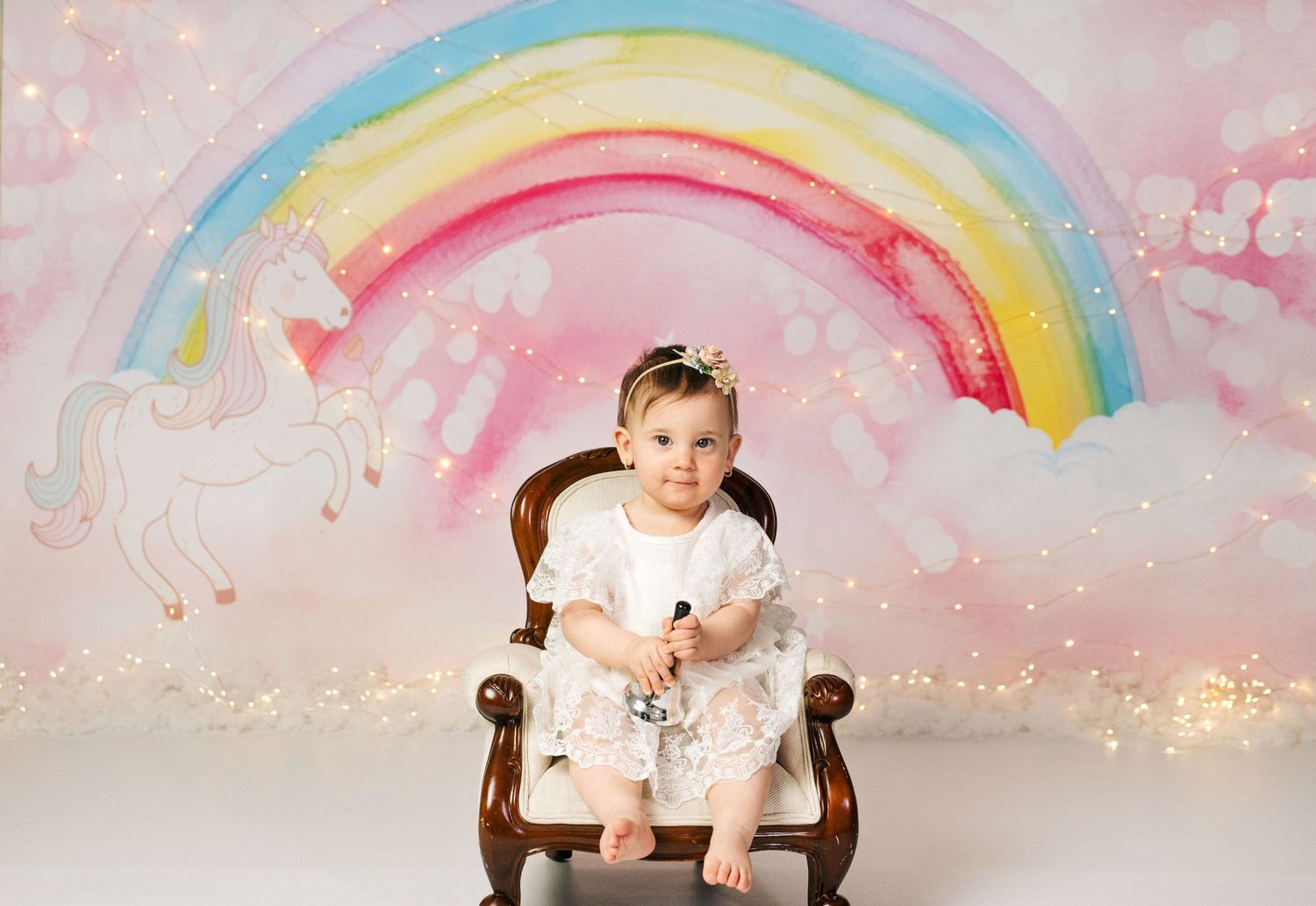 Baby girl in white dress sits on vintage chair before a whimsical rainbow unicorn backdrop with twinkling lights.