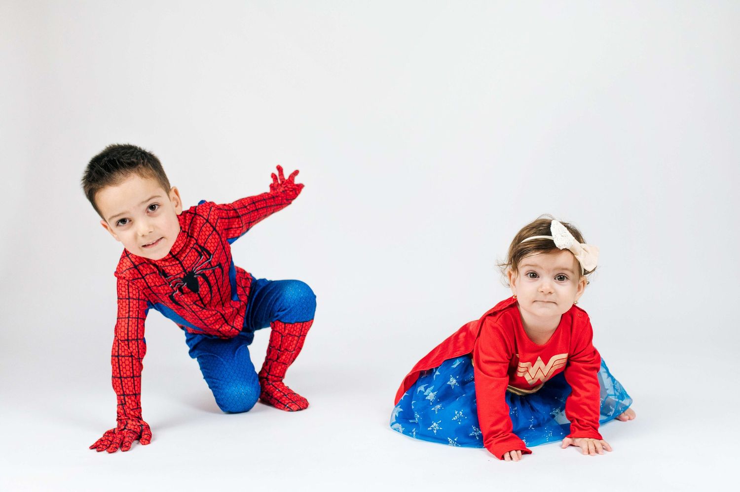 Two children in Spider-Man costumes pose playfully against a white background, one boy and one girl in matching red and blue outfits.