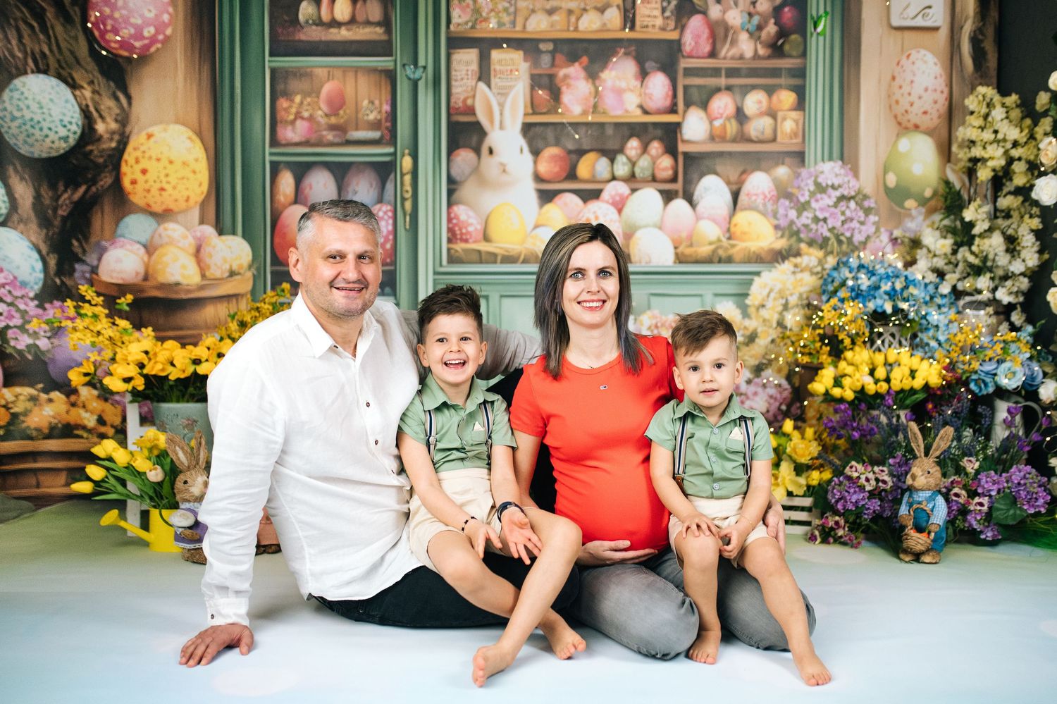 Family of four posing for Easter-themed photo with colorful eggs, flowers, and holiday decorations in background.