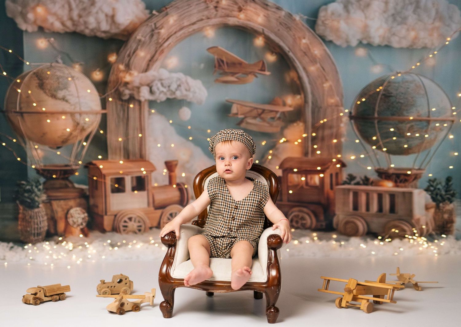 Baby in knit romper sits in vintage chair surrounded by wooden toys, fairy lights, and hot air balloon backdrop.