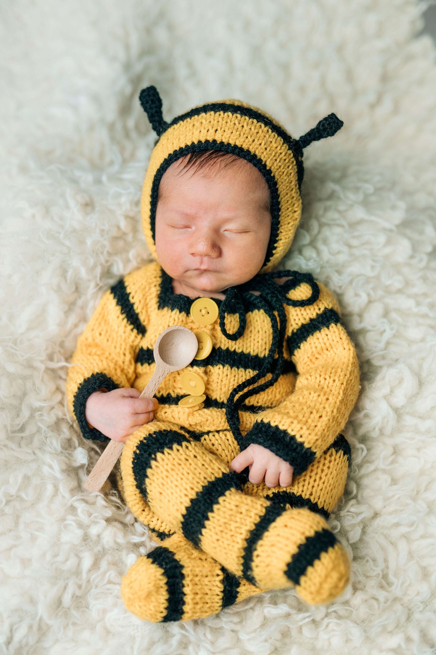 Sleeping newborn baby dressed in a yellow and black knitted bumblebee costume holding a tiny wooden spoon.