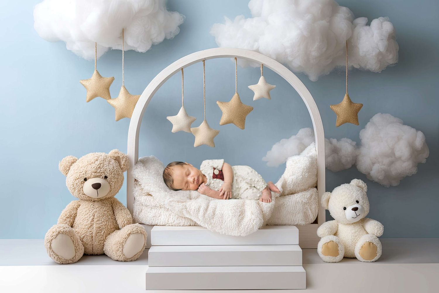 Sleeping newborn baby in white arch crib with teddy bears, star mobiles, and fluffy clouds on blue nursery backdrop.