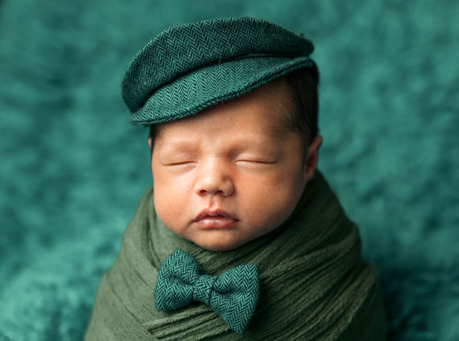 Sleeping newborn baby wrapped in green cloth wearing a green flat cap and bow tie.