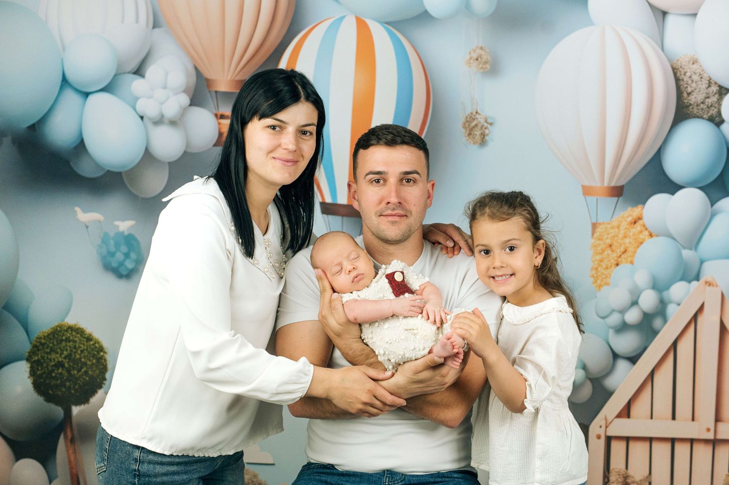 Happy family of four posing with newborn baby in front of colorful hot air balloon backdrop.